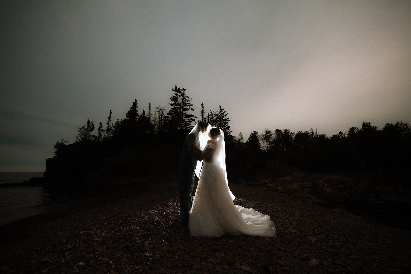 A bride in silhouette standing alone in a dark field, her dress catching the last light
