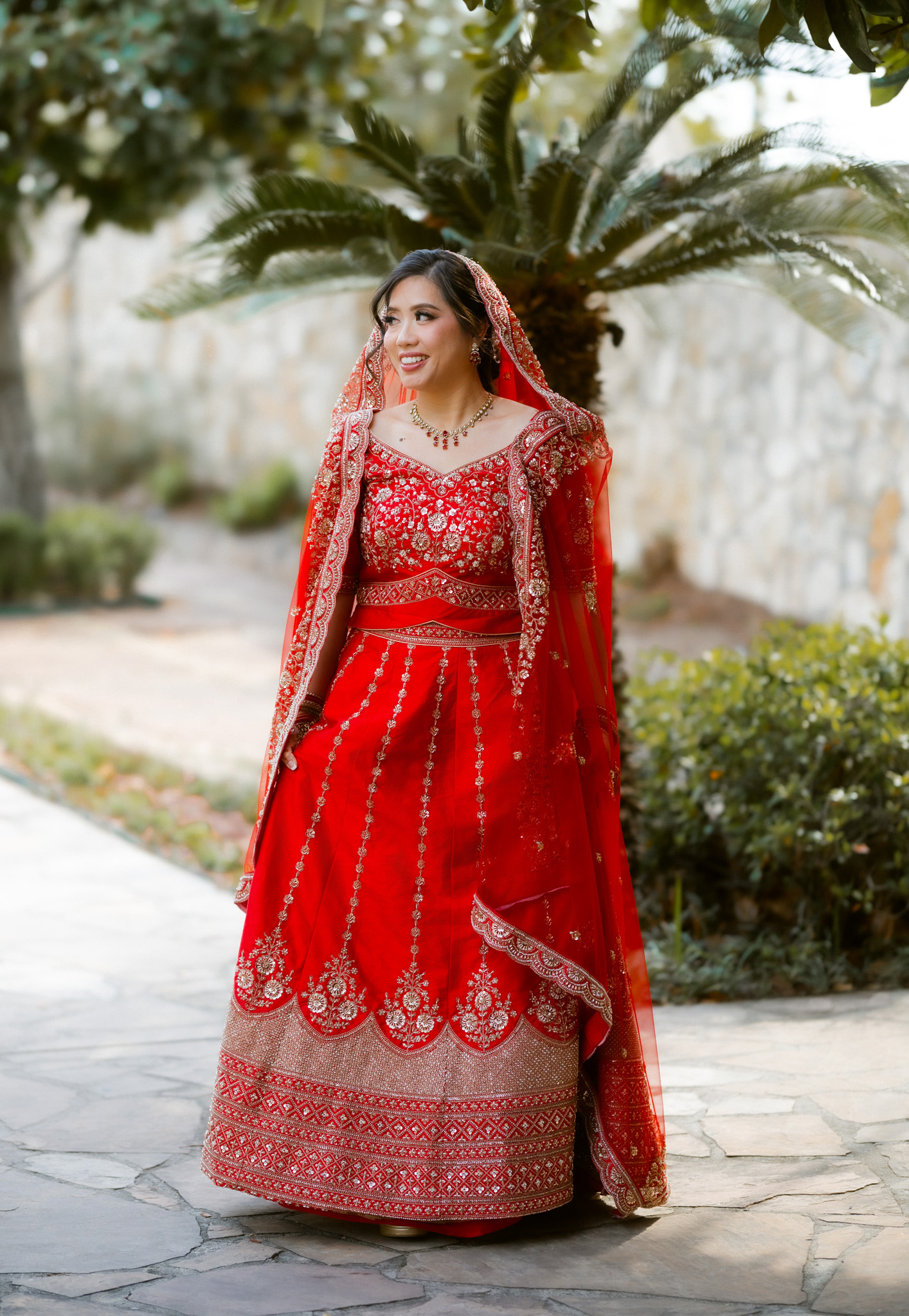 A bride in a red and gold lehenga standing among the palm trees