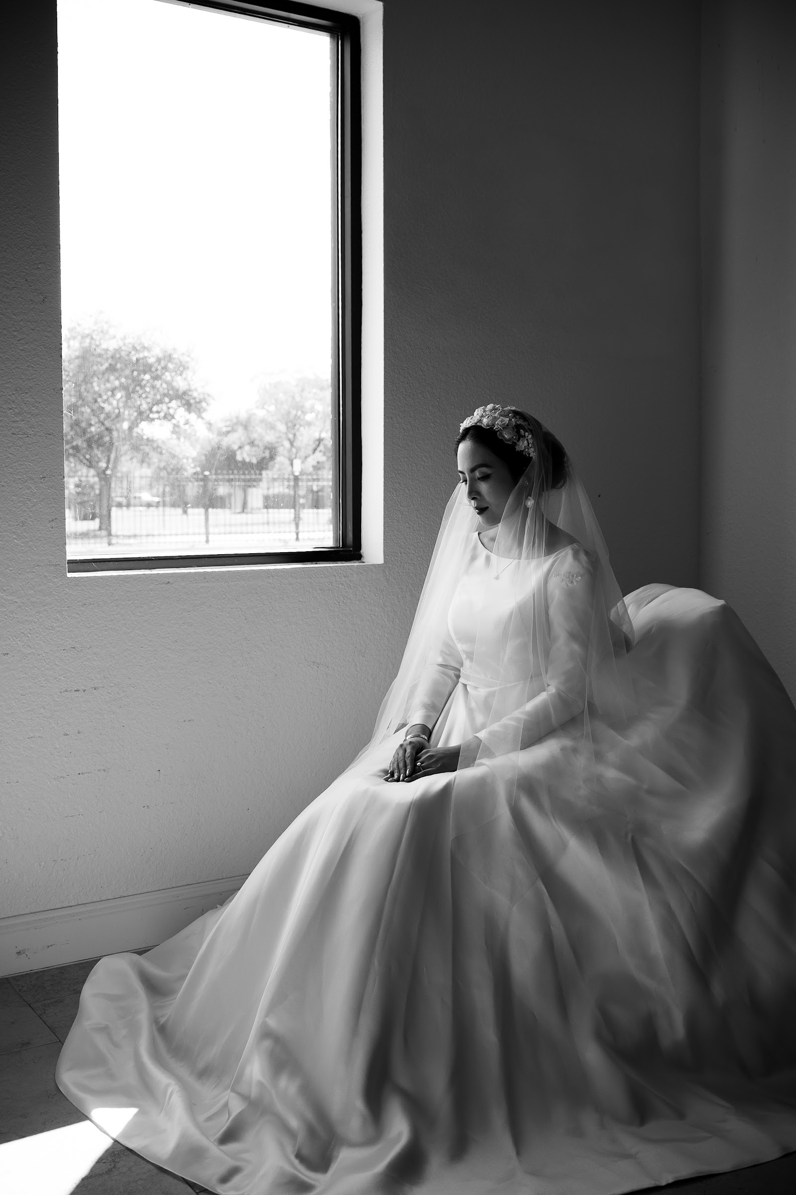 A black-and-white portrait of a bride seated by a tall window, lost in thought